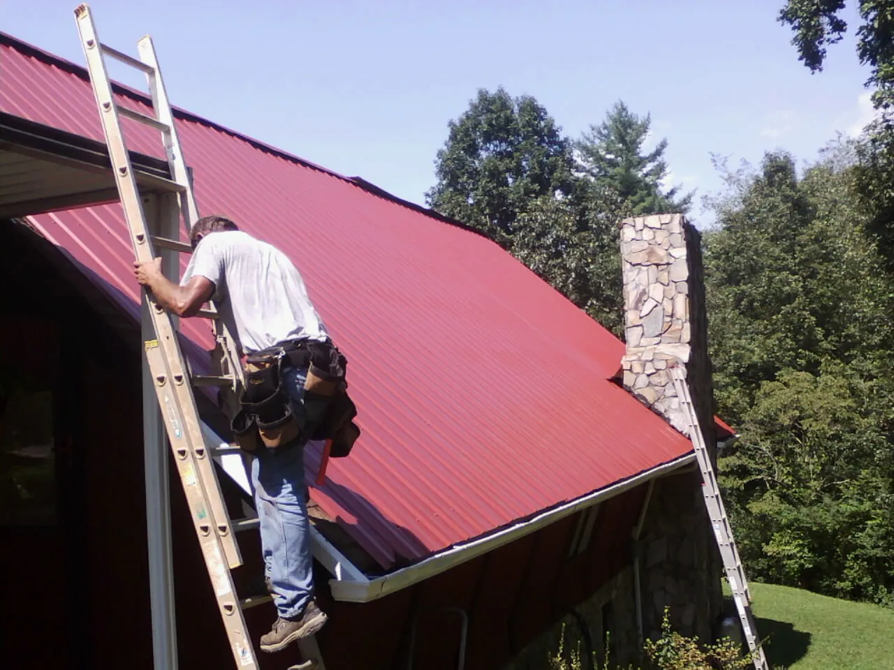 Roofer on a ladder beside a red metal roof and stone chimney — Soffit & Fascia Repair by Roofing Brothers in Alamosa